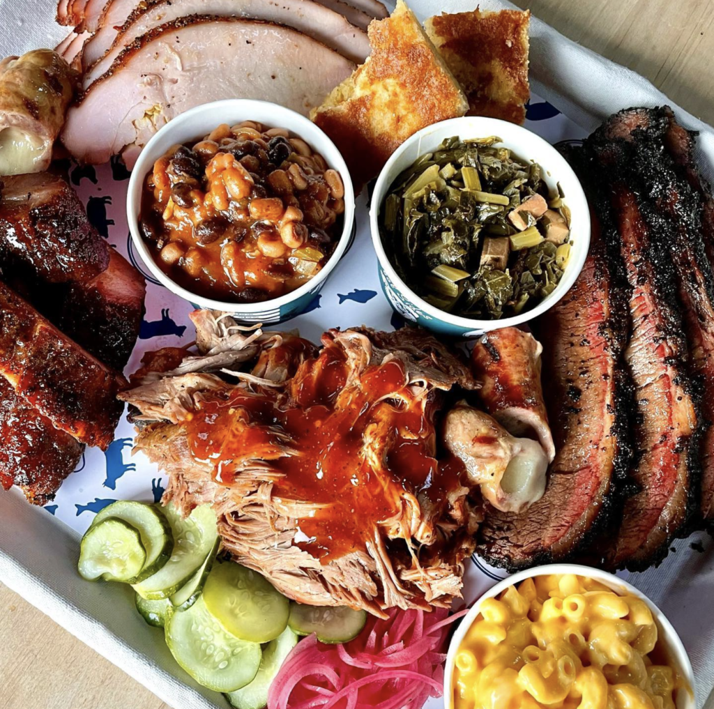 Overhead close-up of a large BBQ platter featuring pulled pork, brisket, turkey, ribs, mac and cheese, collard greens, baked beans, cornbread, and pickles