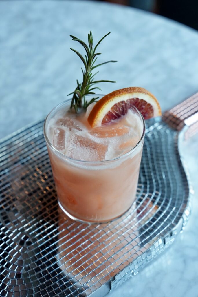 Close-up of a pink cocktail garnished with an orange slice and rosemary.